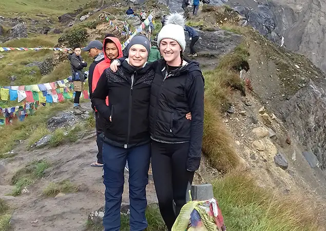 Two women in hiking jackets and beanies stand with their arms around each other on a narrow dirt path. Colorful prayer flags line the trail behind them, and other hikers are visible in the background against a steep mountain slope in the short Annapurna Base Camp Trek.