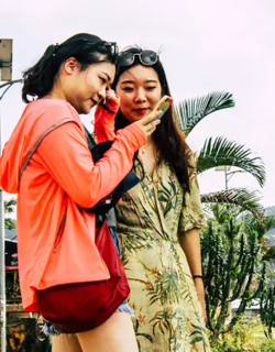 Two women standing outdoors near a cafe, looking at a smartphone screen with green hills and tropical plants in the background.