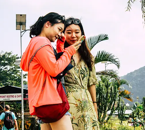 Two women standing outdoors near a cafe, looking at a smartphone screen with green hills and tropical plants in the background.