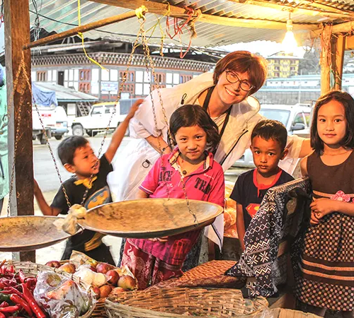 A tourist interacts with Bhutanese children during 5 Days Bhutan Tour at a local market, surrounded by vegetables and spices.
