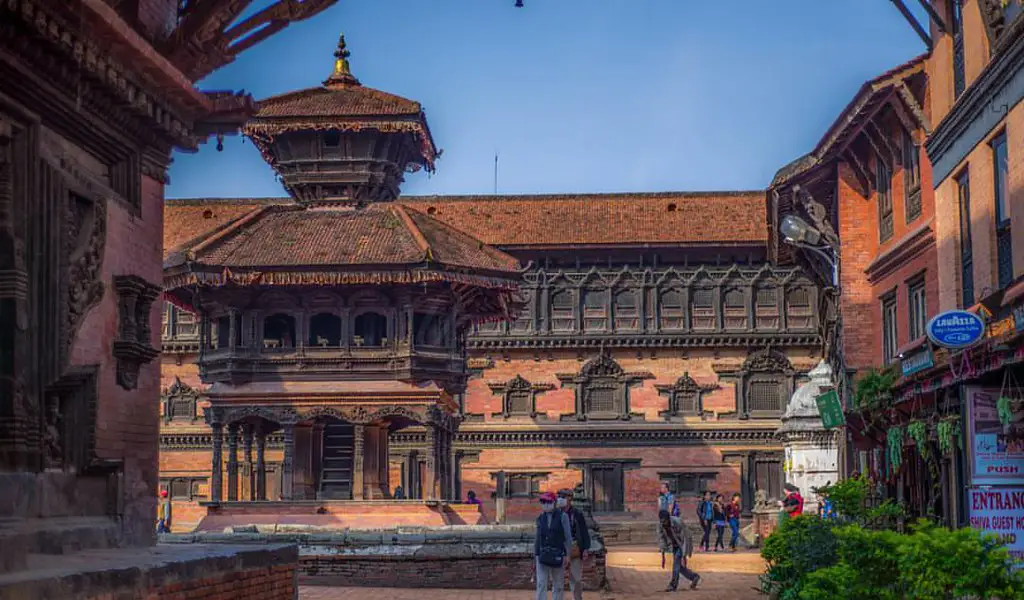 A traditional palace courtyard with carved wooden windows and brick buildings, with people walking through Bhaktapur Durbar Square.