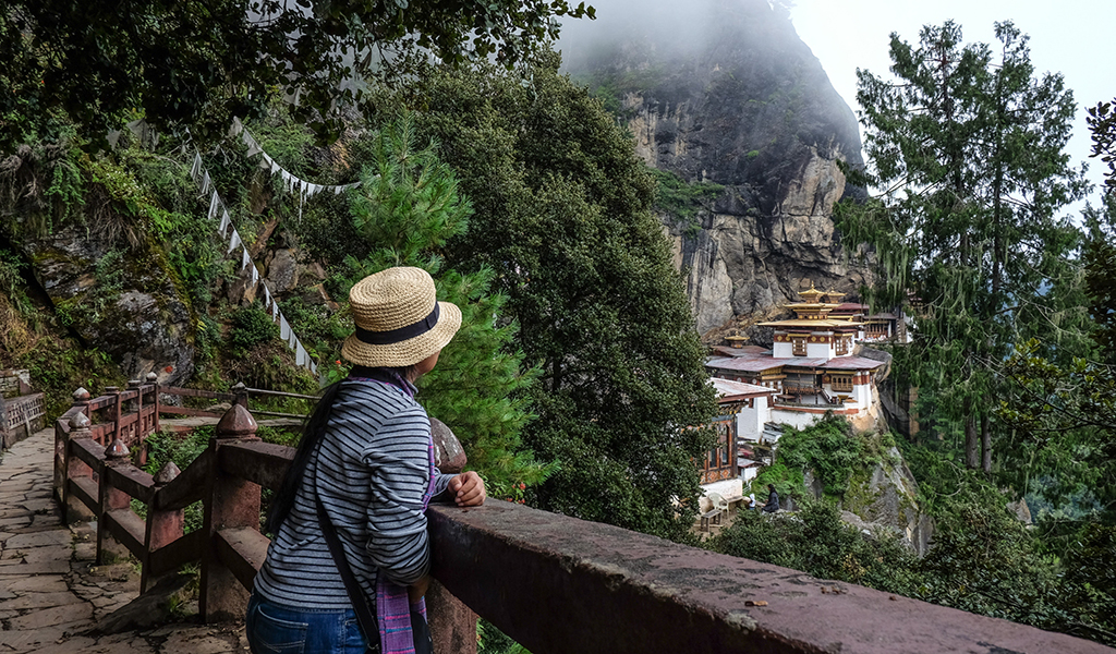 A woman standing on a viewpoint looking toward Taktsang Palphug Monastery (Tiger’s Nest) perched on a cliff amid forested mountains in Paro, Bhutan.