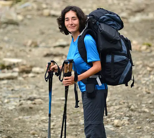 A female trekker wearing a blue shirt carries a large backpack and holds trekking poles while standing on a rocky mountain trail.