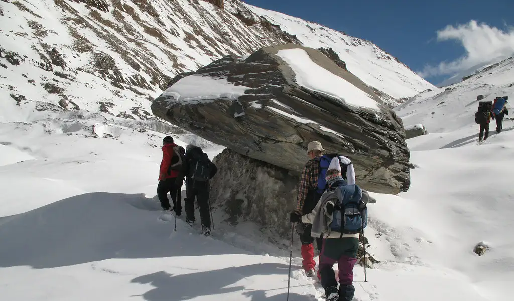 Hikers trek through deep snow beside a massive boulder on the Chhonbardan Glacier, surrounded by snow-covered mountains under a clear blue sky.