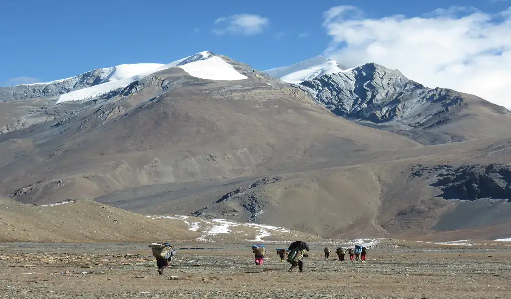 Porters and trekkers crossing a wide alpine valley beneath rugged snow-capped peaks on the Dhaulagiri Trek in Nepal.