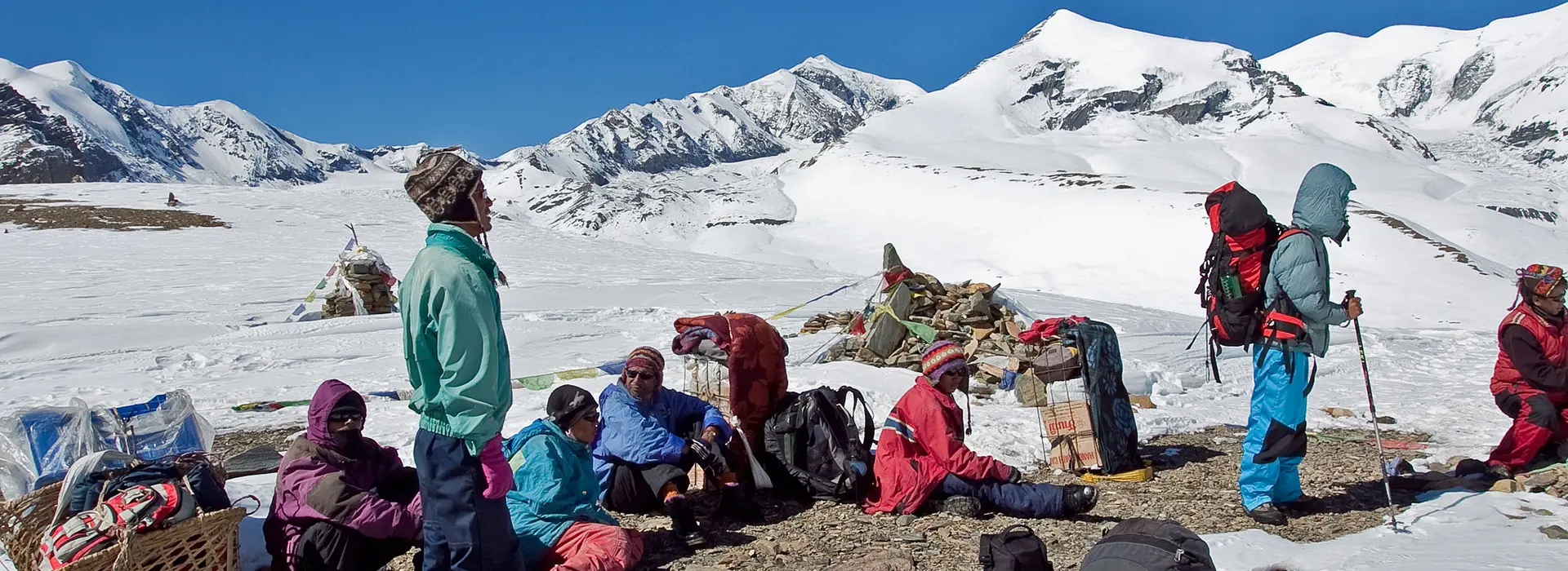 Trekkers resting on a snow-covered high pass with Dhaulagiri and surrounding peaks in the background.