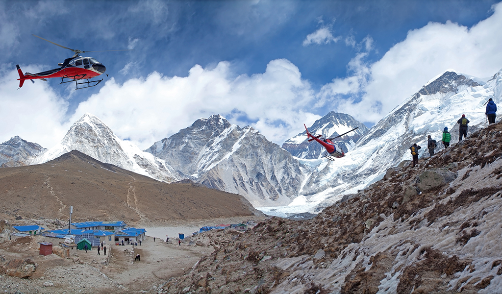 Helicopters flying over Gorak Shep near Everest Base Camp, with trekkers on a rocky slope and snow-covered Himalayan peaks in the background.
