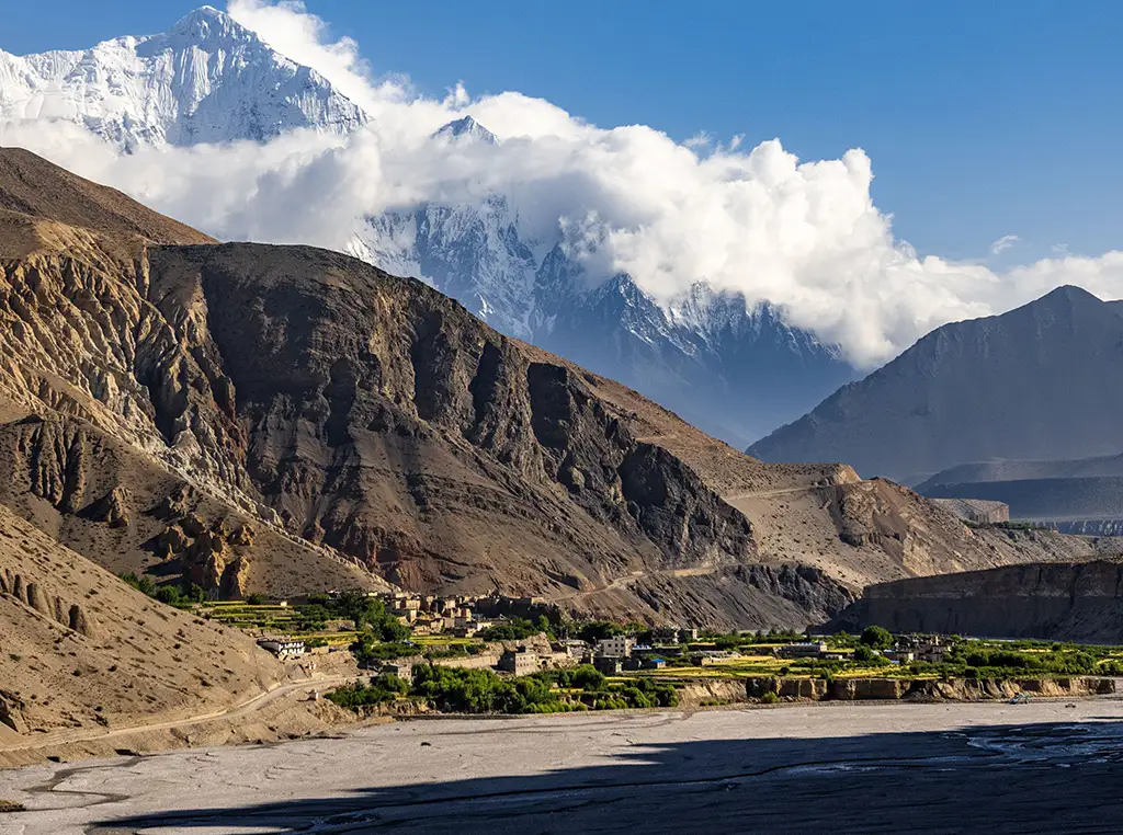 Kali Gandaki River valley in Mustang, Nepal, with a small village and green fields beneath rugged cliffs and snow-capped Himalayan peaks.