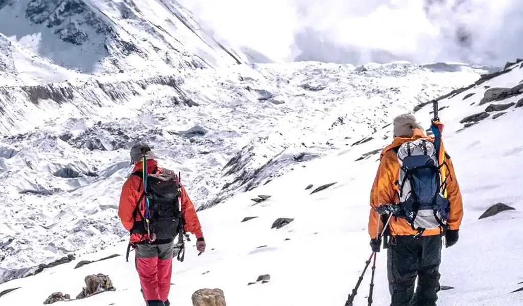 Two trekkers walking across a snow-covered trail near Lobuche Peak, carrying backpacks and trekking poles with glaciers and high Himalayan terrain ahead.