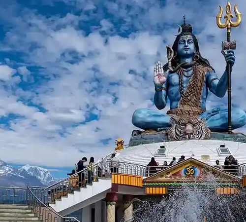 A large blue statue of Lord Shiva sits atop a hill, holding a trident, with mountains and a cloudy sky in the background.