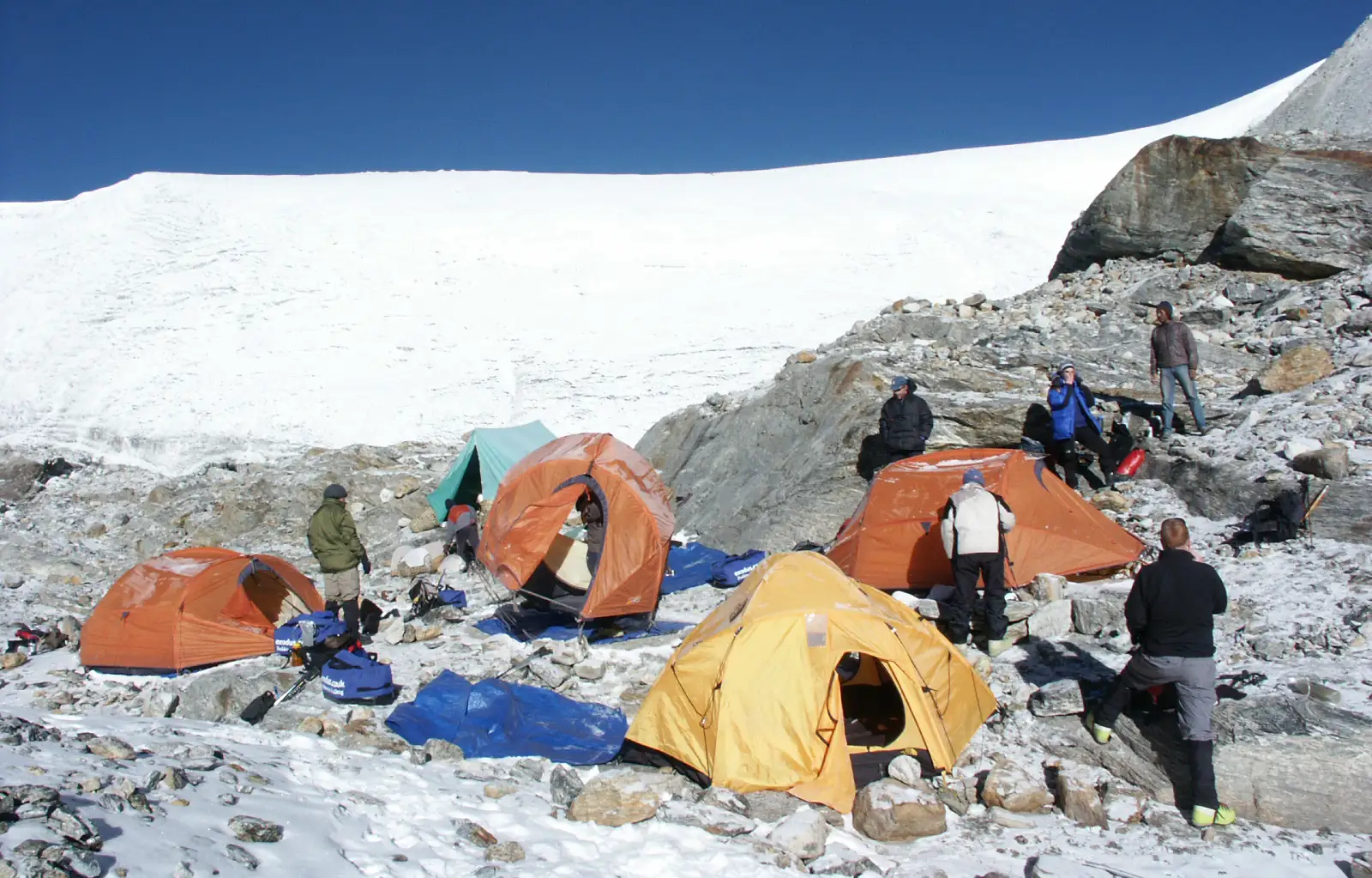 Climbers pack their tents at the snowy Mera La campsite with the glacier and blue sky in the background.