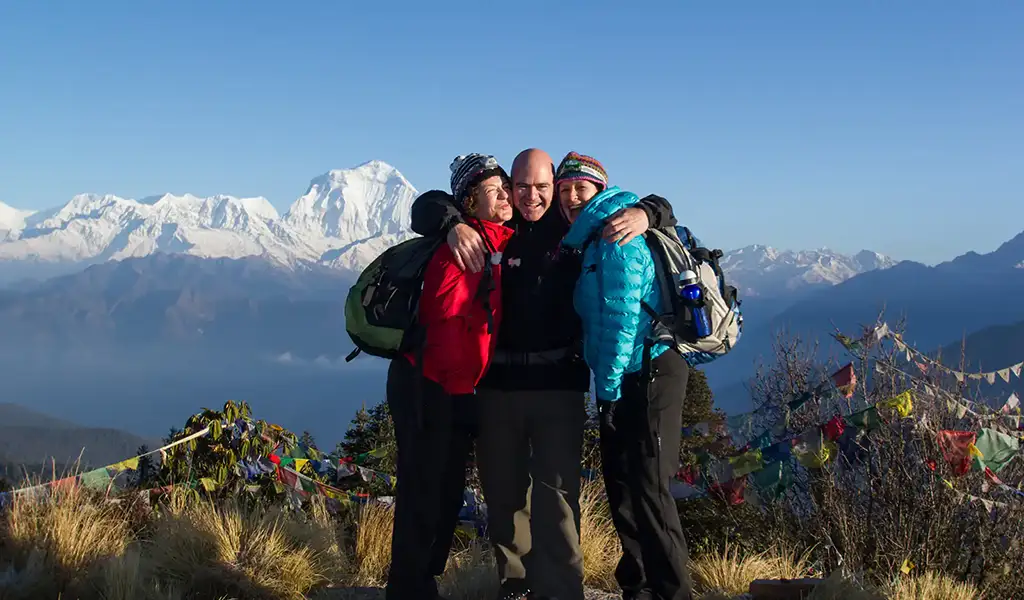 Annapurna base Camp Trek Photo of three hikers hugging at a Himalayan viewpoint with snow-capped peaks and prayer flags behind them.