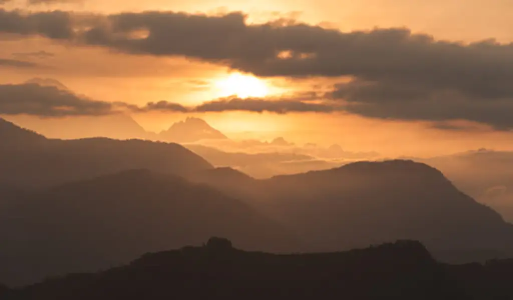 Sunrise over layered hills and distant Himalayan peaks as golden light filters through clouds, viewed from Sarangkot near Pokhara, Nepal.