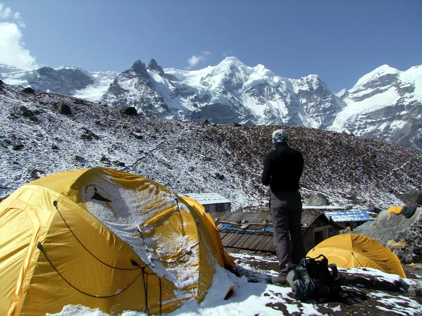 A climber stands beside yellow tents at a snowy campsite in Khare, surrounded by towering Himalayan peaks.