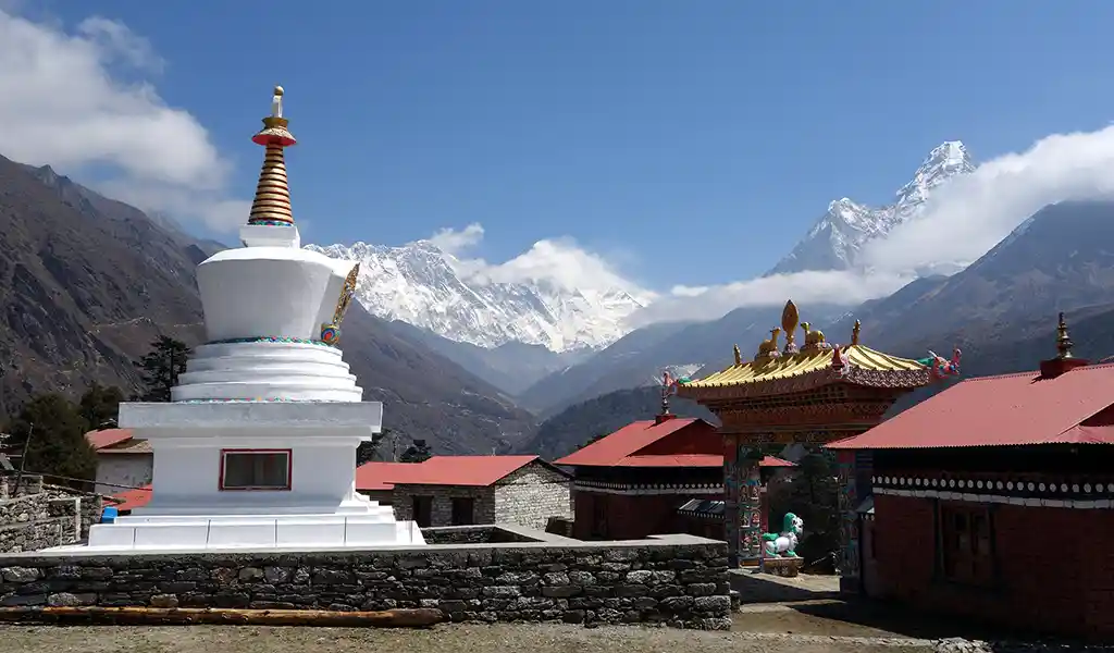 White Buddhist stupa at Tengboche monastery with red-roofed buildings and snow-covered Himalayan peaks, including Everest and Lhotse, in the background.