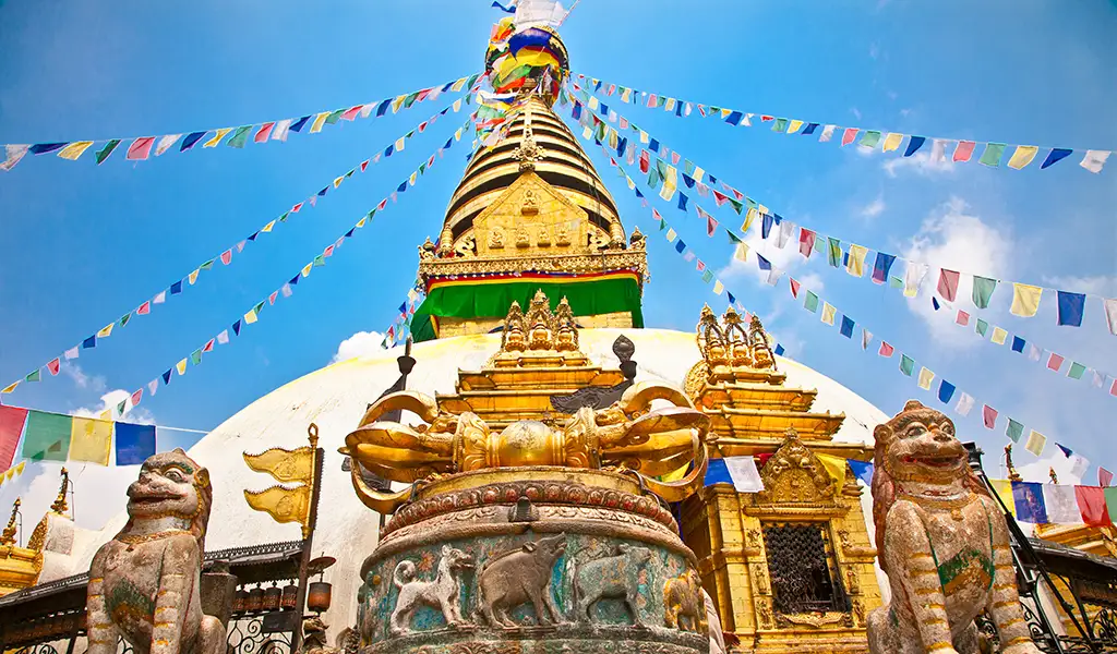 The golden spire and white dome of Swayambhunath Stupa in Kathmandu are decorated with colorful prayer flags under a blue sky, with stone guardian statues in the foreground.
