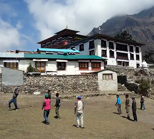 People playing volleyball in an open courtyard near Tengboche Monastery, with monastery buildings and Himalayan mountains in the background.
