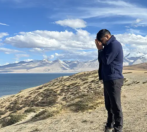 A man stands in prayer facing the calm blue waters of Lake Mansarovar with distant mountain during Mount Kailash tour from Nepal.