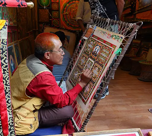 An artist sits indoors painting a detailed traditional artwork on a framed canvas, surrounded by colorful designs and tools.