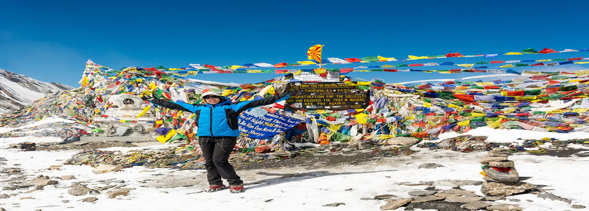A trekker standing at Thorong La Pass with arms raised, surrounded by colorful prayer flags and snow-covered terrain on the Annapurna Circuit in Nepal.