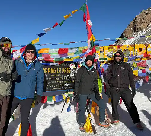 Group of trekkers standing at Thorong La Pass on the Annapurna Circuit, surrounded by snow, prayer flags, and a sign marking the high mountain pass.