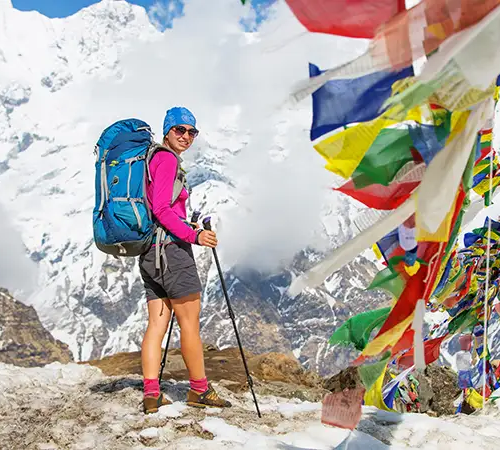 Trekker standing beside colorful prayer flags on a high mountain trail during the Annapurna Circuit trek, with snow-covered Himalayan peaks in the background.