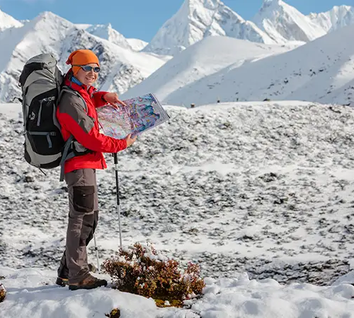 A hiker wearing an orange beanie and red jacket stands in snowy mountains holding a map, with a large backpack and trekking poles.