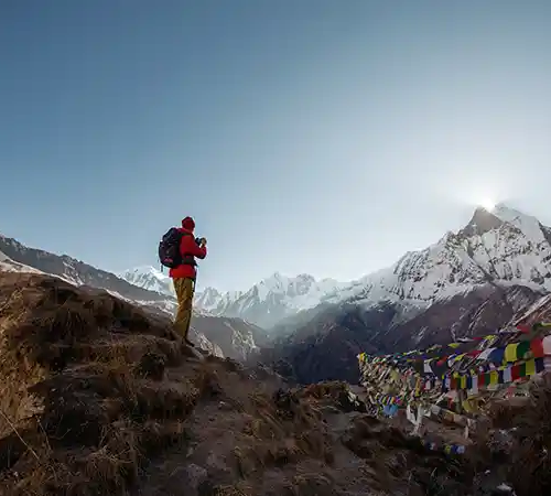 Trekker standing on a narrow mountain trail with prayer flags and snow-covered Himalayan peaks in the distance, showing annapurna circuit trek height and high-altitude terrain.