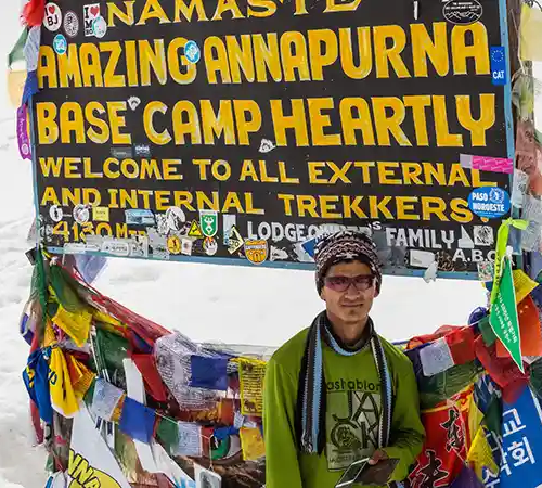 A trekker stands smiling in front of the Annapurna Base Camp welcome sign, surrounded by colorful prayer flags and snow.