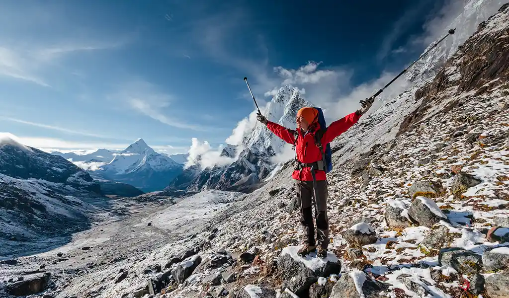 Trekker standing on a rocky, snow-covered slope with arms raised and trekking poles, surrounded by Himalayan peaks, showing conditions during the best time for Everest Base Camp trek.