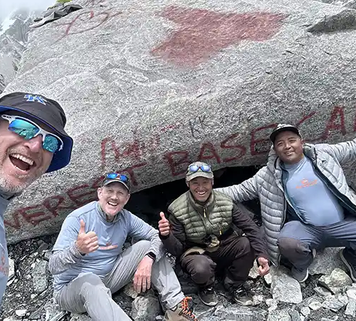 Group of trekkers posing beside a rock marked “Everest Base Camp,” surrounded by rocky terrain and high-altitude mountain surroundings.