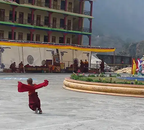 A young Buddhist monk in red robes runs with arms outstretched across an open courtyard at a monastery complex.