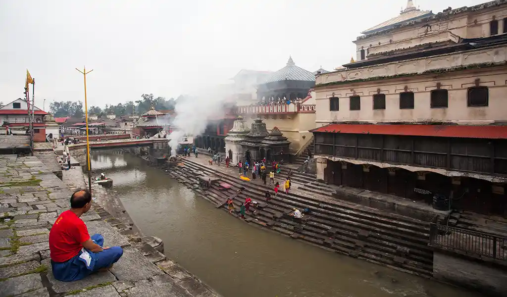 Cremation ghats along the Bagmati River at Pashupatinath Temple in Kathmandu, Nepal, with smoke rising and visitors on the steps.