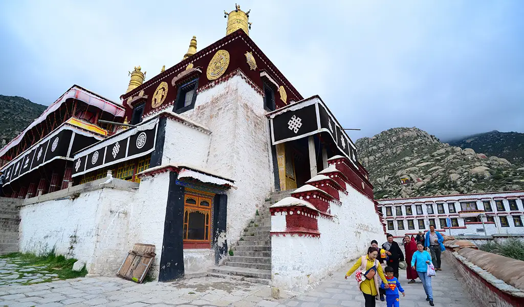 Exterior of Drepung Monastery in Tibet with white and maroon walls, golden roof ornaments, and visitors walking beside the monastery buildings.