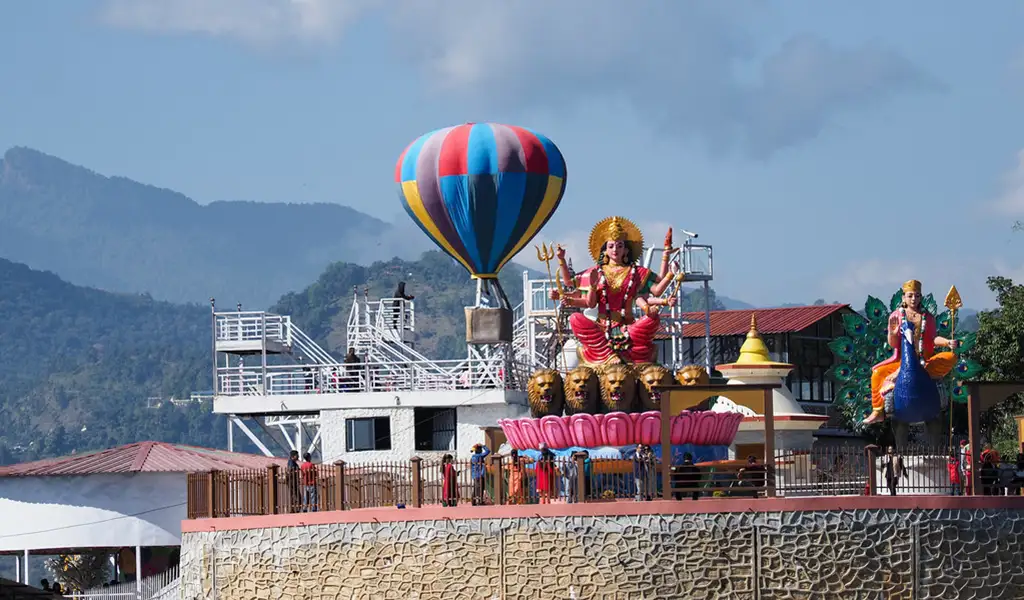 A large statue of Goddess Durga seated on a lotus pedestal is displayed at the Pumdikot temple complex in Pokhara, with a colorful hot air balloon structure and hills in the background.