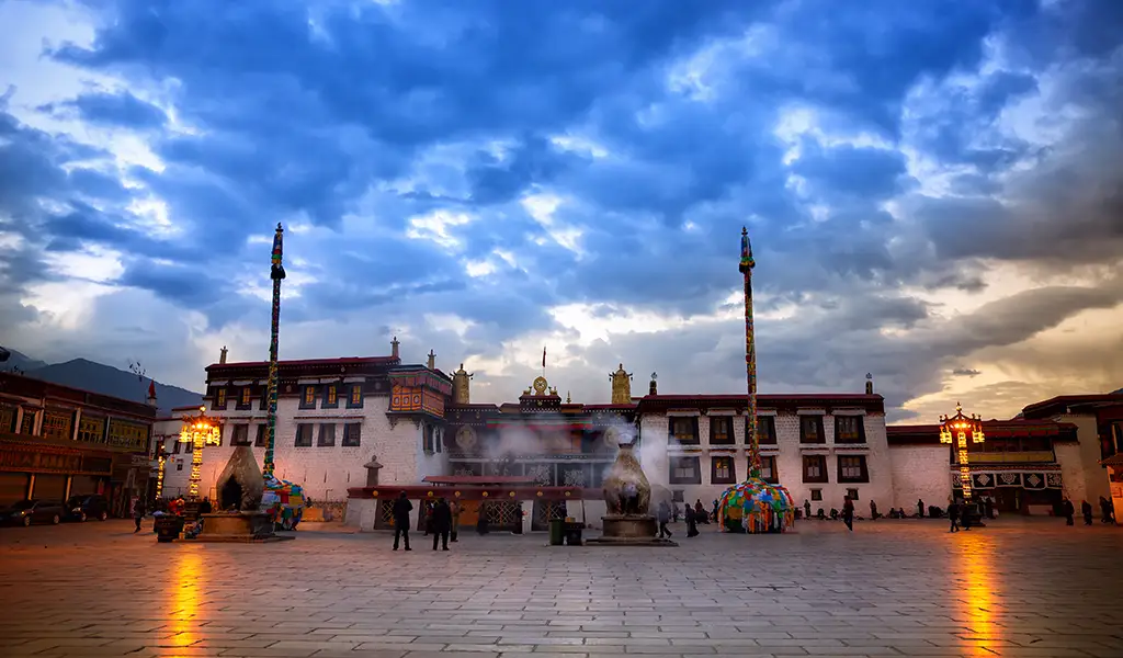 Devotees gather in the courtyard of Jokhang Temple in Lhasa, Tibet, with incense smoke rising under a dramatic blue evening sky.