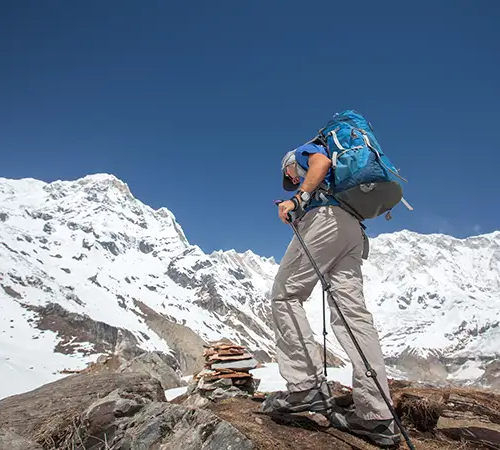 Trekker climbing a rocky section of the Annapurna Circuit trail with a backpack and trekking poles, surrounded by snow-covered Himalayan peaks.