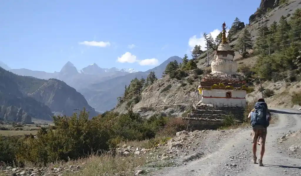 Trekker walking along a mountain trail past a small Buddhist stupa, with distant Himalayan peaks visible on the route, showing the Annapurna Base Camp trek distance.