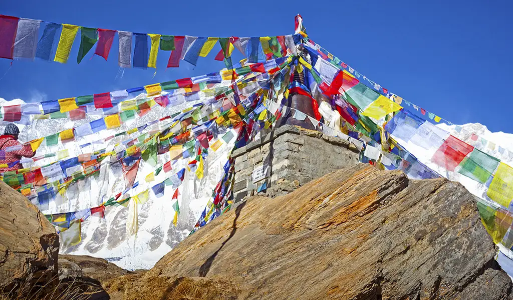 A stone chorten covered with colorful prayer flags on a high mountain trail, marking the Annapurna Circuit trek distance with snow-covered Himalayan peaks behind.