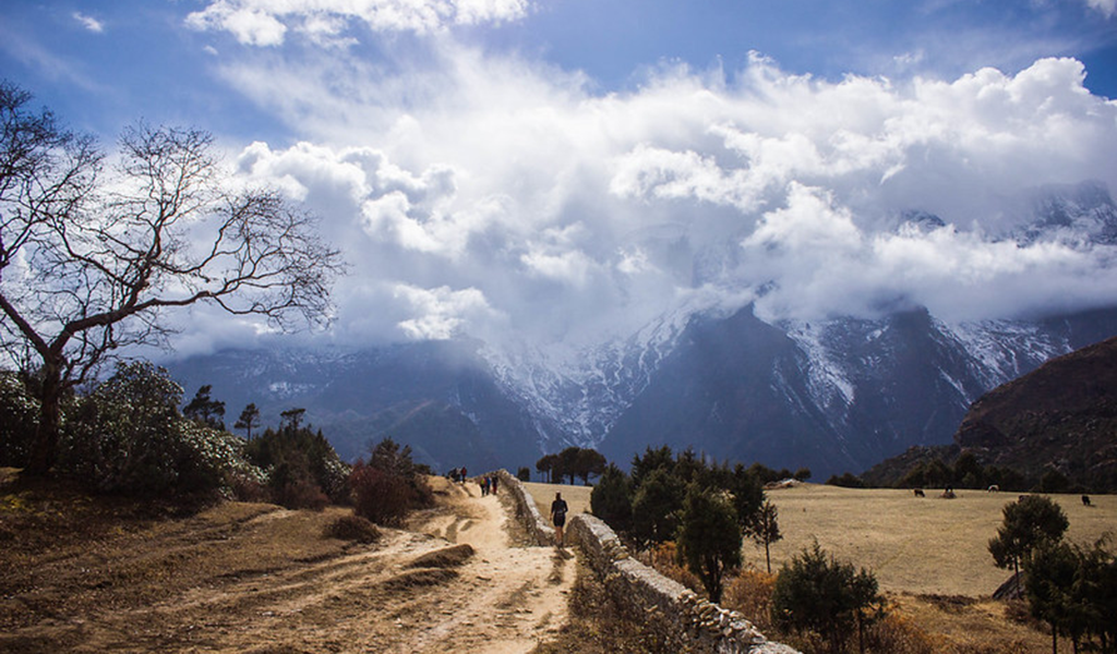 A mountain trail winding through open fields toward snow-covered Himalayan peaks under dramatic clouds, often seen while planning everest base camp trek cost and route conditions.