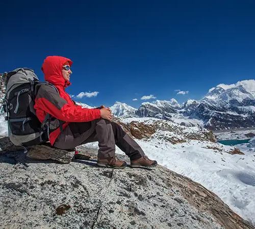 Trekker sitting on a rocky outcrop with a backpack, resting and looking out over snow-covered Himalayan peaks near the Everest Base Camp route.