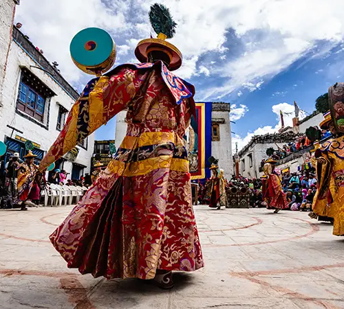 Performers in ornate traditional costumes dance in a courtyard during a Himalayan cultural festival.