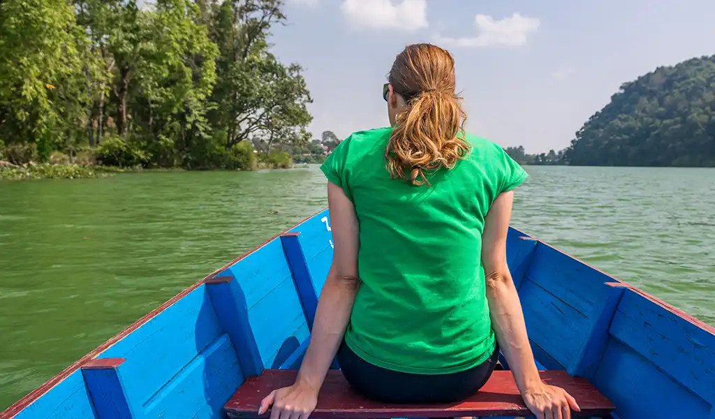A woman sits in a wooden boat on Phewa Lake in Pokhara, Nepal, with green hills and trees along the shoreline.