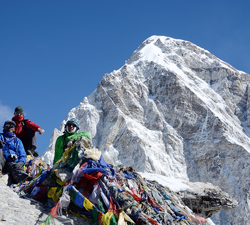 Trekkers sitting near prayer flags at the base of Kala Patthar, with a steep snow-covered Himalayan peak rising behind them near Gorakshep.
