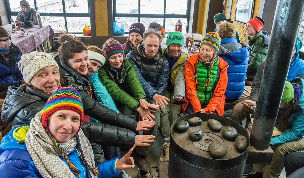 A group of trekkers in warm jackets and hats sits inside a mountain lodge, warming their hands around a metal stove during the Annapurna Circuit Trek.
