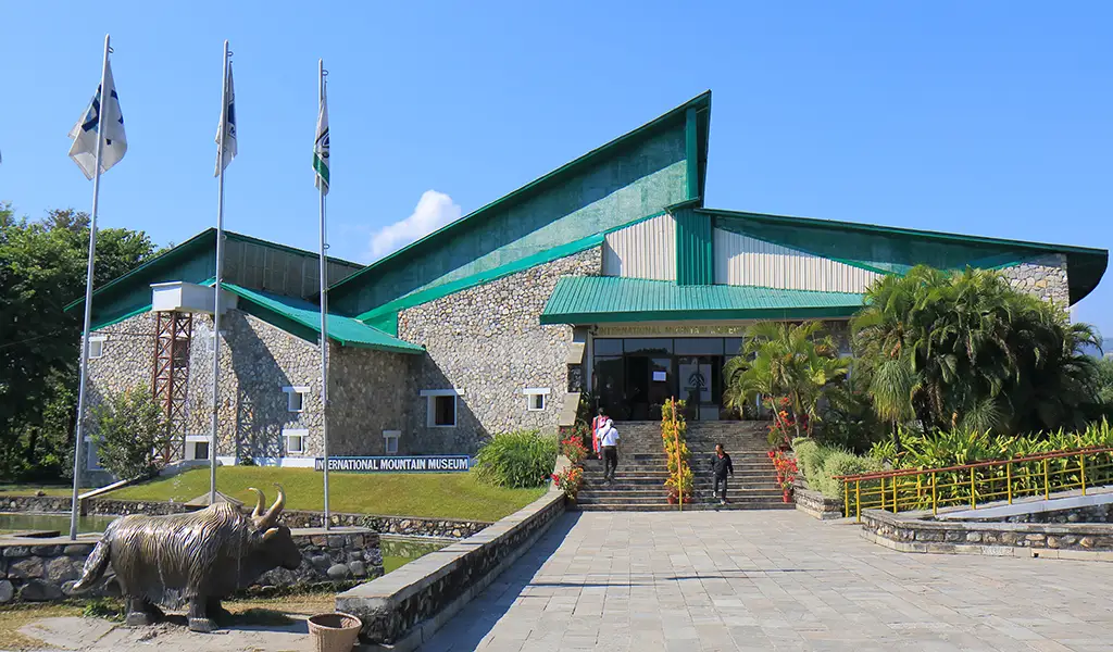 Exterior view of the International Mountain Museum in Pokhara, Nepal, with a modern stone building, green roof, flags, and a yak statue near the entrance.
