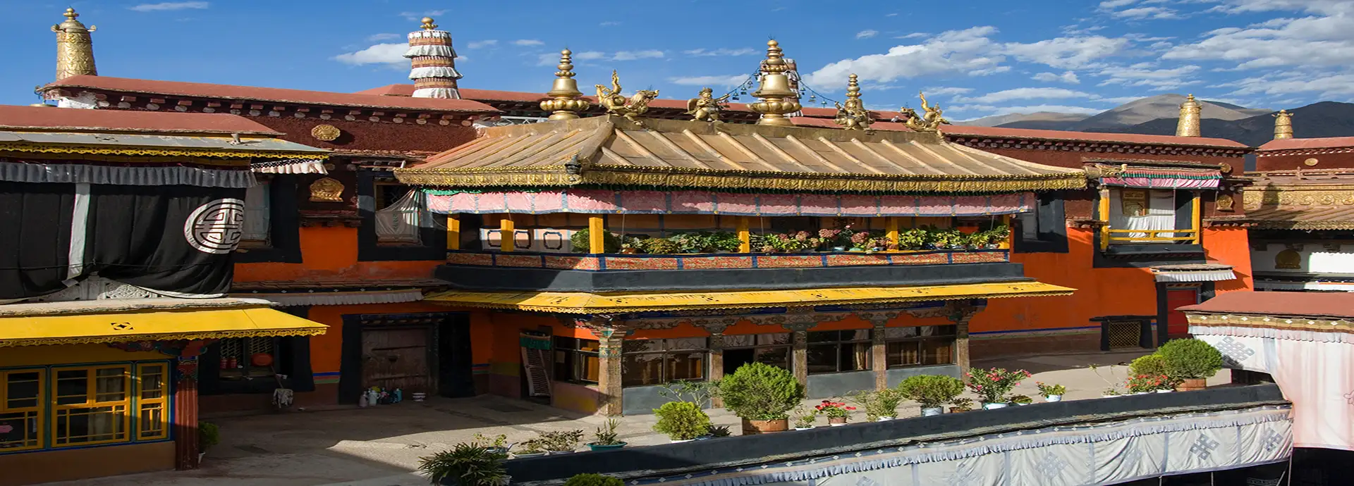 Ornate Tibetan Buddhist temple with golden roofs, colorful walls, prayer flags, and mountain scenery under a blue sky.
