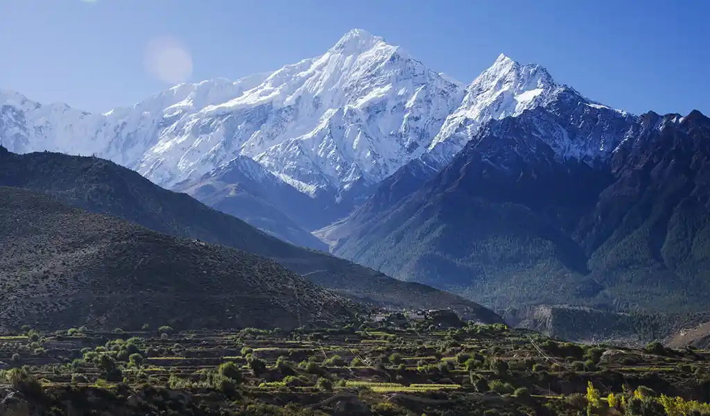 Snow-capped Nilgiri and surrounding Himalayan peaks rise above the Jomsom valley in Mustang, Nepal, with terraced fields and hills in the foreground.