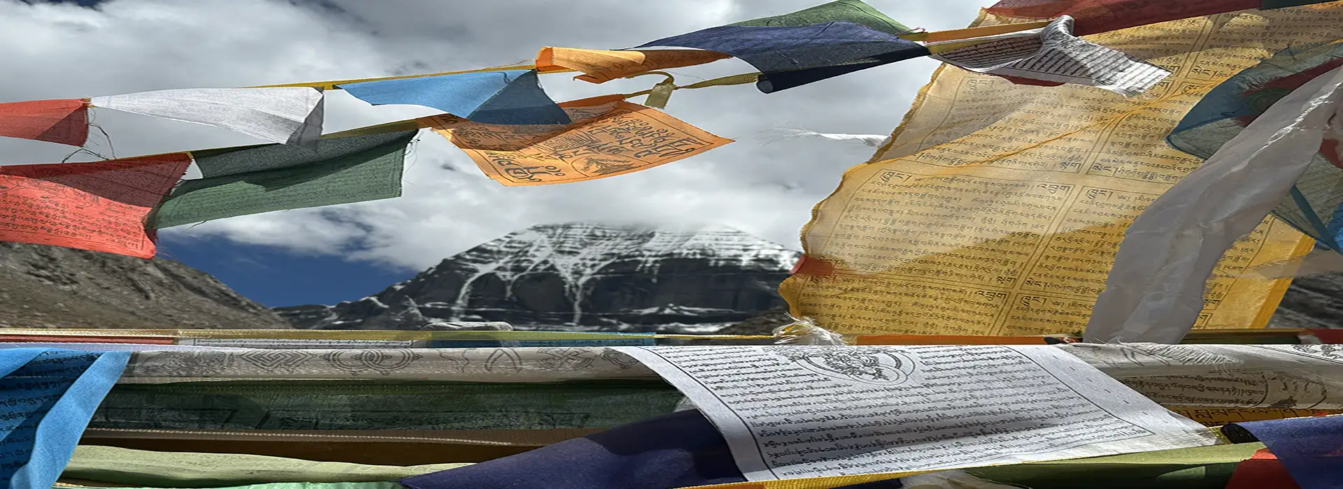 Colorful Tibetan prayer flags fluttering in the wind with Mount Kailash rising in the background under a dramatic sky.