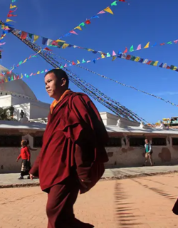 Two Buddhist monks in maroon robes walk past Boudhanath Stupa, with colorful prayer flags overhead under a clear blue sky.